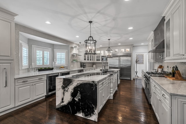 kitchen with wine cooler, high end appliances, hanging light fixtures, a kitchen island with sink, and wall chimney range hood