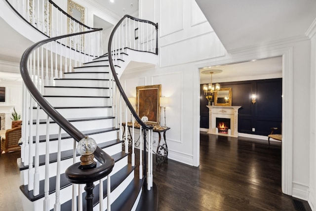 staircase with wood finished floors, a high ceiling, a lit fireplace, crown molding, and a decorative wall