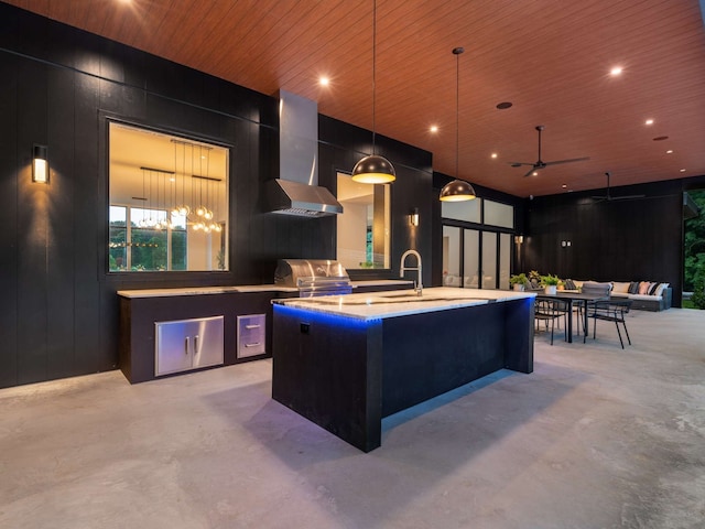 kitchen featuring pendant lighting, wall chimney exhaust hood, ceiling fan, an island with sink, and wood ceiling