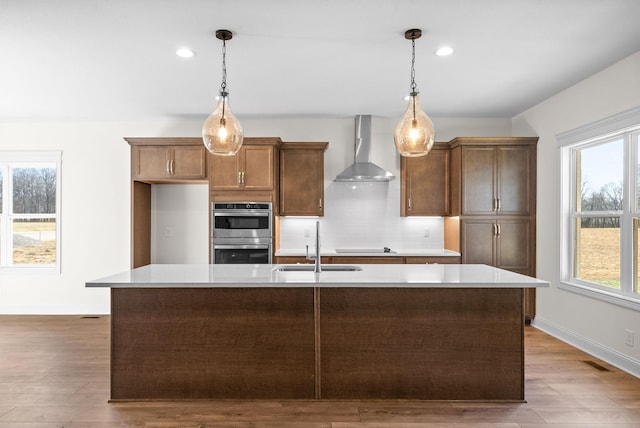 kitchen featuring pendant lighting, stainless steel double oven, a kitchen island with sink, and wall chimney exhaust hood