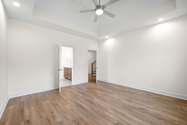 unfurnished bedroom with light wood-type flooring, a tray ceiling, ensuite bath, and ceiling fan