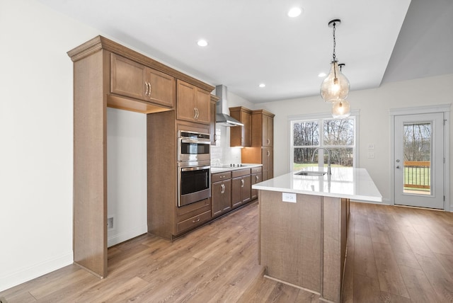 kitchen featuring a center island with sink, wall chimney range hood, sink, decorative light fixtures, and stainless steel double oven