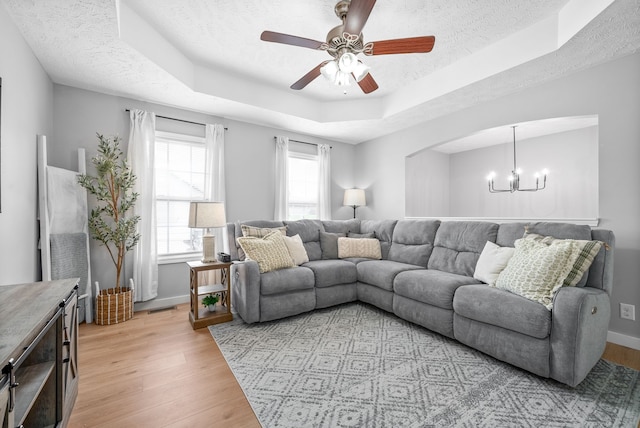 living room with ceiling fan with notable chandelier, a textured ceiling, light wood-type flooring, and a raised ceiling