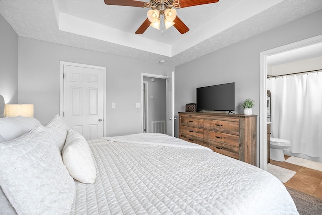 bedroom featuring a textured ceiling, a tray ceiling, ensuite bath, and ceiling fan