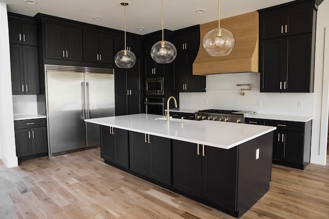 kitchen with pendant lighting, built in appliances, light hardwood / wood-style floors, and a center island with sink