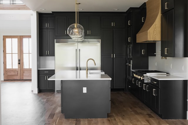 kitchen with french doors, custom range hood, pendant lighting, hardwood / wood-style flooring, and a center island with sink