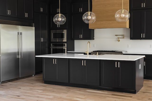 kitchen with built in appliances, a kitchen island with sink, decorative light fixtures, and light wood-type flooring