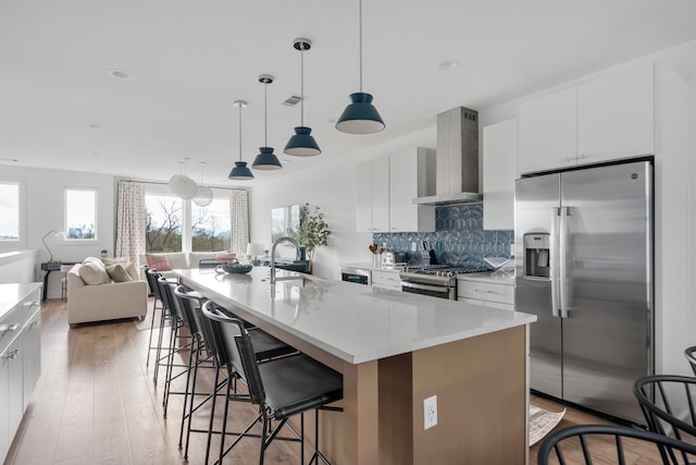 kitchen featuring a center island with sink, white cabinets, stainless steel appliances, and wall chimney range hood