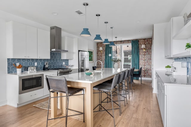 kitchen with white cabinetry, wall chimney exhaust hood, hanging light fixtures, stainless steel appliances, and an island with sink