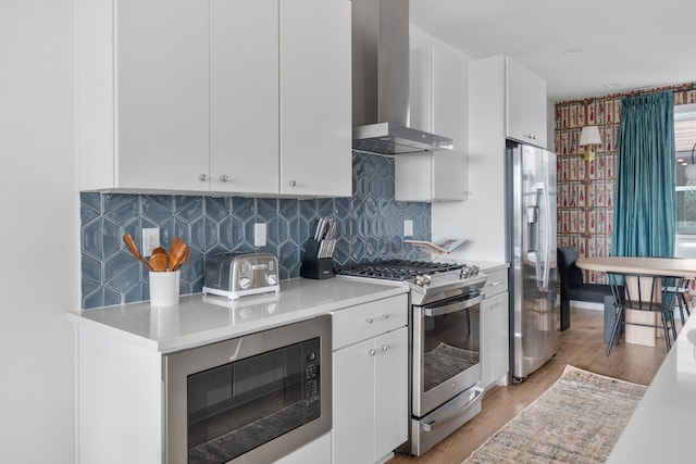 kitchen featuring white cabinetry, stainless steel appliances, wall chimney range hood, decorative backsplash, and light wood-type flooring