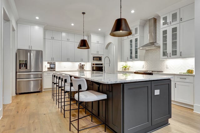 kitchen featuring white cabinets, wall chimney range hood, hanging light fixtures, an island with sink, and stainless steel appliances