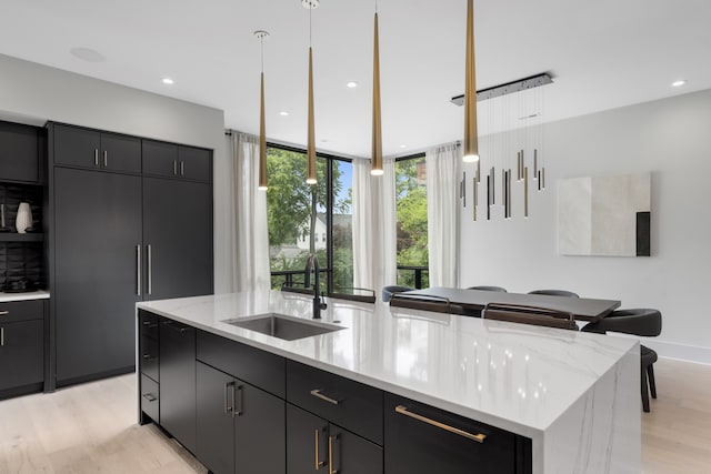 kitchen featuring light stone countertops, light wood-type flooring, sink, pendant lighting, and an island with sink