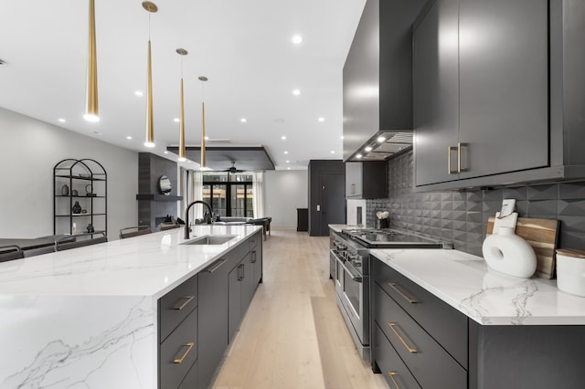 kitchen with pendant lighting, a large island with sink, wall chimney exhaust hood, high end stove, and light stone counters