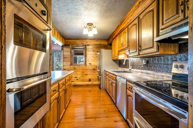 kitchen featuring wood walls, ventilation hood, sink, light wood-type flooring, and appliances with stainless steel finishes