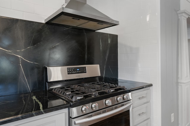 kitchen featuring ornate columns, stainless steel gas range oven, dark stone counters, wall chimney range hood, and white cabinetry