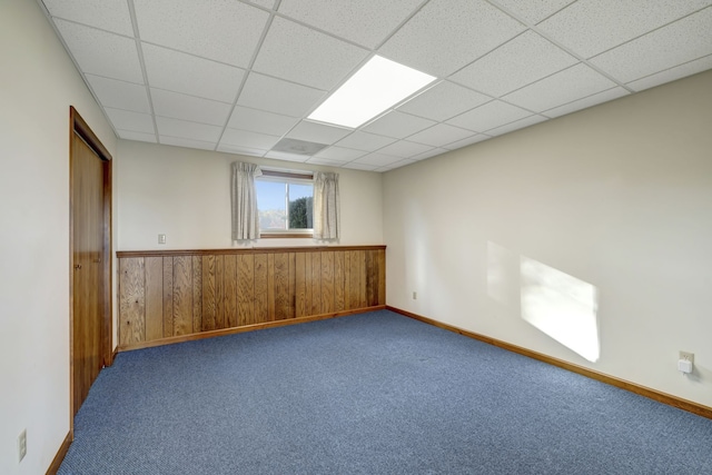 carpeted empty room featuring wood walls, a drop ceiling, and wainscoting