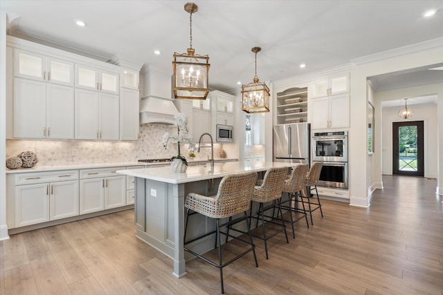 kitchen featuring appliances with stainless steel finishes, custom exhaust hood, a center island with sink, white cabinets, and light hardwood / wood-style floors