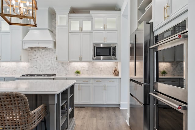 kitchen with light wood-type flooring, custom exhaust hood, stainless steel appliances, decorative light fixtures, and white cabinets