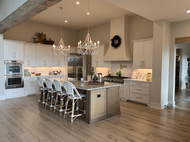 kitchen with light wood-type flooring, a kitchen island with sink, white cabinets, custom range hood, and appliances with stainless steel finishes