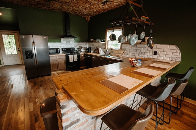 kitchen featuring kitchen peninsula, decorative backsplash, wall chimney exhaust hood, black appliances, and hardwood / wood-style flooring