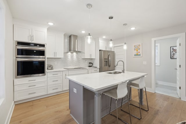 kitchen featuring white cabinets, appliances with stainless steel finishes, wall chimney exhaust hood, sink, and a kitchen island with sink