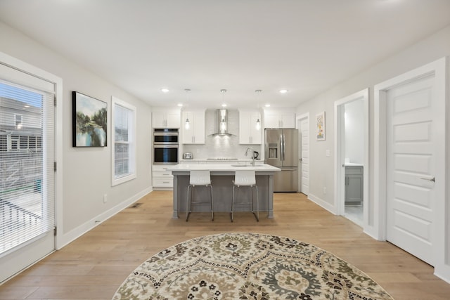 kitchen with hanging light fixtures, white cabinets, stainless steel appliances, a center island with sink, and wall chimney exhaust hood