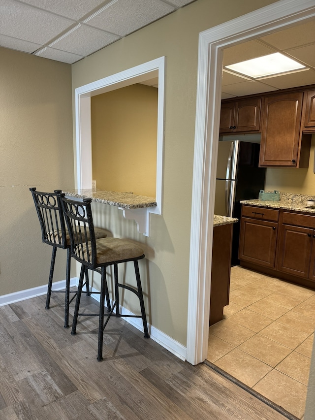 kitchen featuring a drop ceiling, stainless steel fridge, light stone countertops, light wood-type flooring, and a kitchen bar