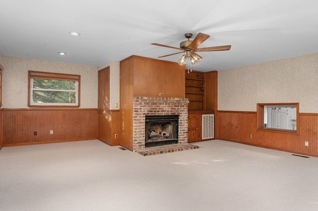 unfurnished living room with built in shelves, light colored carpet, ceiling fan, a fireplace, and wood walls