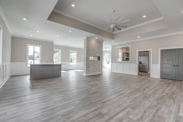 unfurnished living room with ornamental molding, a raised ceiling, ceiling fan, light hardwood / wood-style flooring, and a stone fireplace