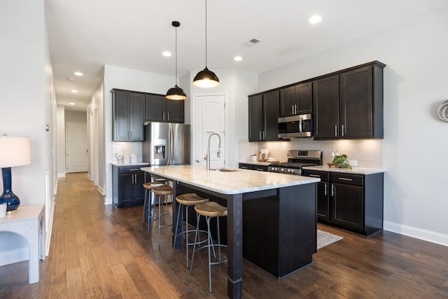 kitchen with a kitchen island with sink, dark wood-type flooring, sink, appliances with stainless steel finishes, and decorative light fixtures