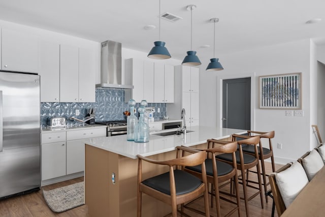 kitchen featuring white cabinets, wall chimney range hood, hanging light fixtures, an island with sink, and stainless steel appliances