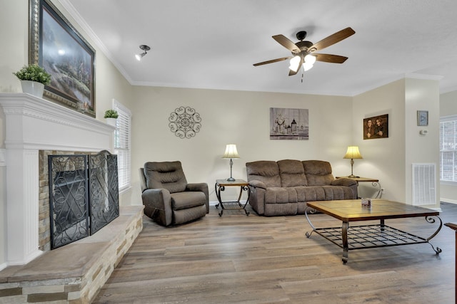 living room featuring hardwood / wood-style flooring, ceiling fan, a stone fireplace, and crown molding