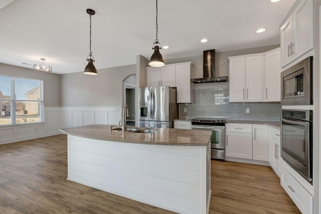 kitchen featuring appliances with stainless steel finishes, hanging light fixtures, wall chimney range hood, and a center island with sink