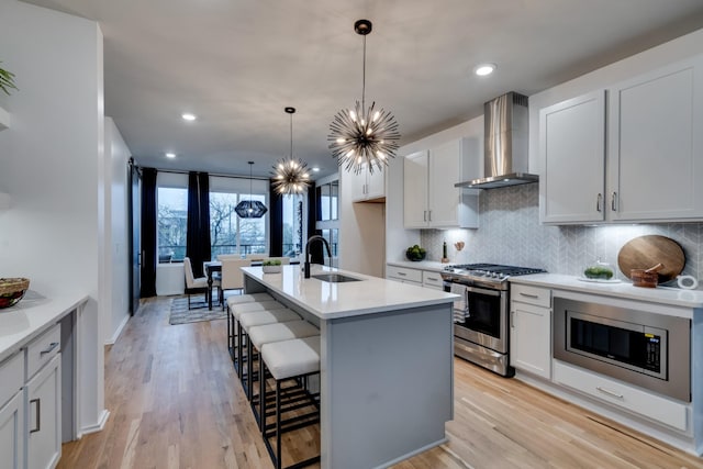 kitchen with a kitchen island with sink, wall chimney range hood, appliances with stainless steel finishes, light hardwood / wood-style floors, and white cabinetry