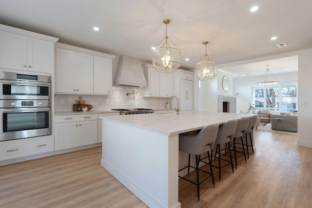 kitchen featuring white cabinets, custom range hood, stainless steel double oven, and an island with sink