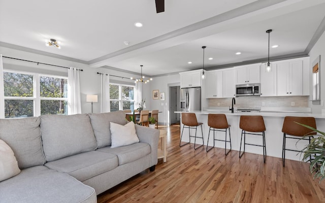 living room featuring ceiling fan with notable chandelier, sink, light wood-type flooring, ornamental molding, and beam ceiling