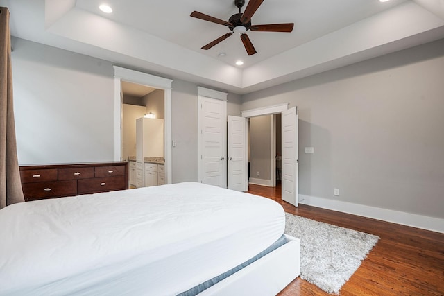 bedroom with a raised ceiling, ceiling fan, ensuite bathroom, and wood-type flooring