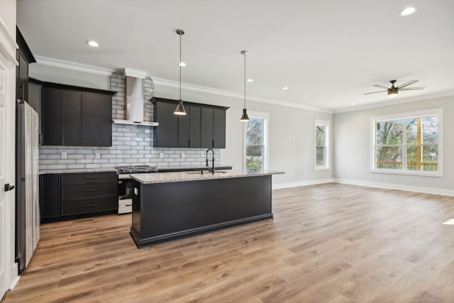 kitchen with wall chimney range hood, light hardwood / wood-style flooring, decorative light fixtures, a kitchen island with sink, and appliances with stainless steel finishes