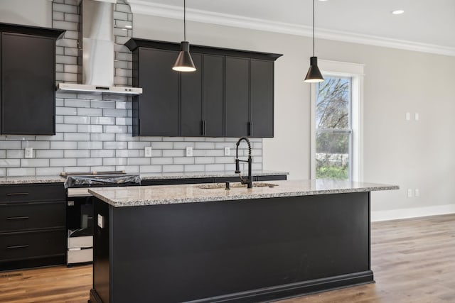 kitchen featuring wall chimney range hood, stainless steel range oven, pendant lighting, a kitchen island with sink, and light wood-type flooring