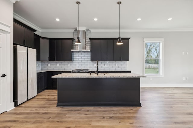 kitchen with a center island with sink, wall chimney range hood, hanging light fixtures, and stainless steel refrigerator