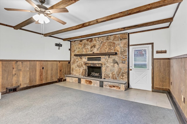 unfurnished living room with a stone fireplace, wooden walls, ceiling fan, beamed ceiling, and light colored carpet