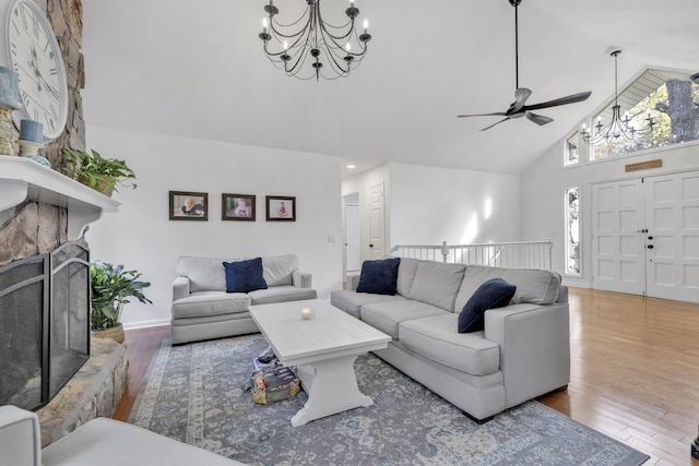 living room featuring hardwood / wood-style floors, ceiling fan with notable chandelier, high vaulted ceiling, and a stone fireplace