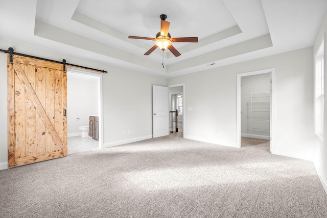 unfurnished bedroom featuring ensuite bath, a raised ceiling, ceiling fan, a barn door, and a spacious closet