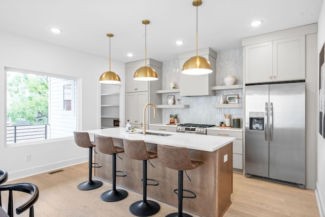 kitchen featuring white cabinets, light wood-type flooring, an island with sink, decorative light fixtures, and stainless steel appliances