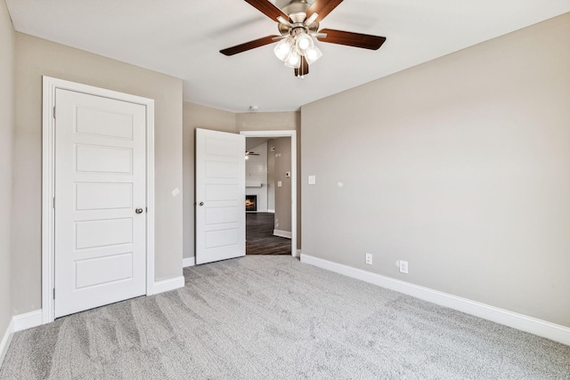 unfurnished bedroom featuring a ceiling fan, baseboards, a lit fireplace, and carpet flooring