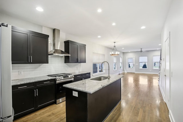 kitchen with wall chimney range hood, sink, stainless steel gas range, light wood-type flooring, and an island with sink