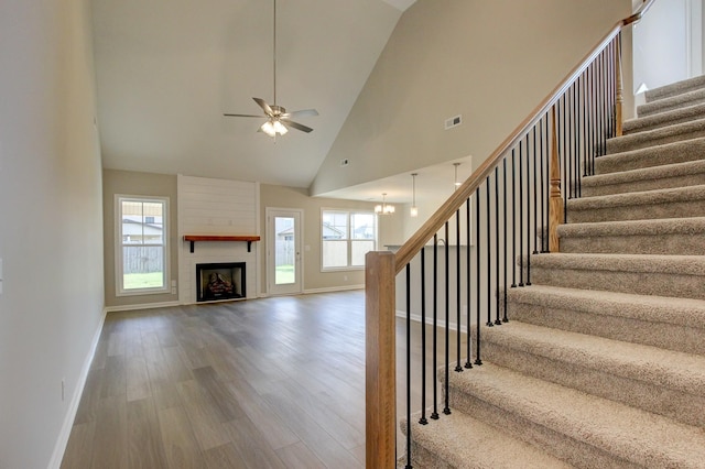 staircase with hardwood / wood-style floors, ceiling fan with notable chandelier, a large fireplace, and high vaulted ceiling