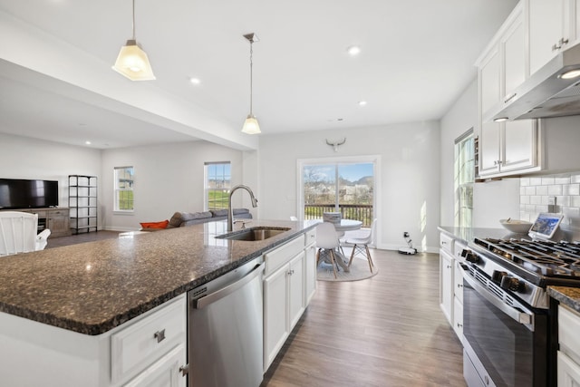 kitchen featuring stainless steel appliances, a kitchen island with sink, sink, dark stone countertops, and hanging light fixtures