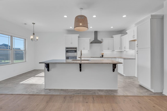 kitchen with a center island with sink, white cabinets, light stone counters, wall chimney range hood, and pendant lighting