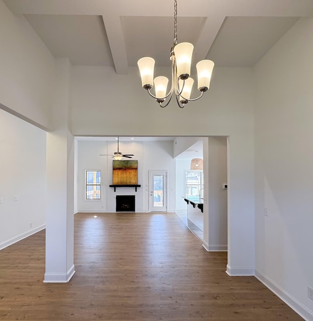 unfurnished living room featuring beam ceiling, a fireplace, wood finished floors, baseboards, and ceiling fan with notable chandelier